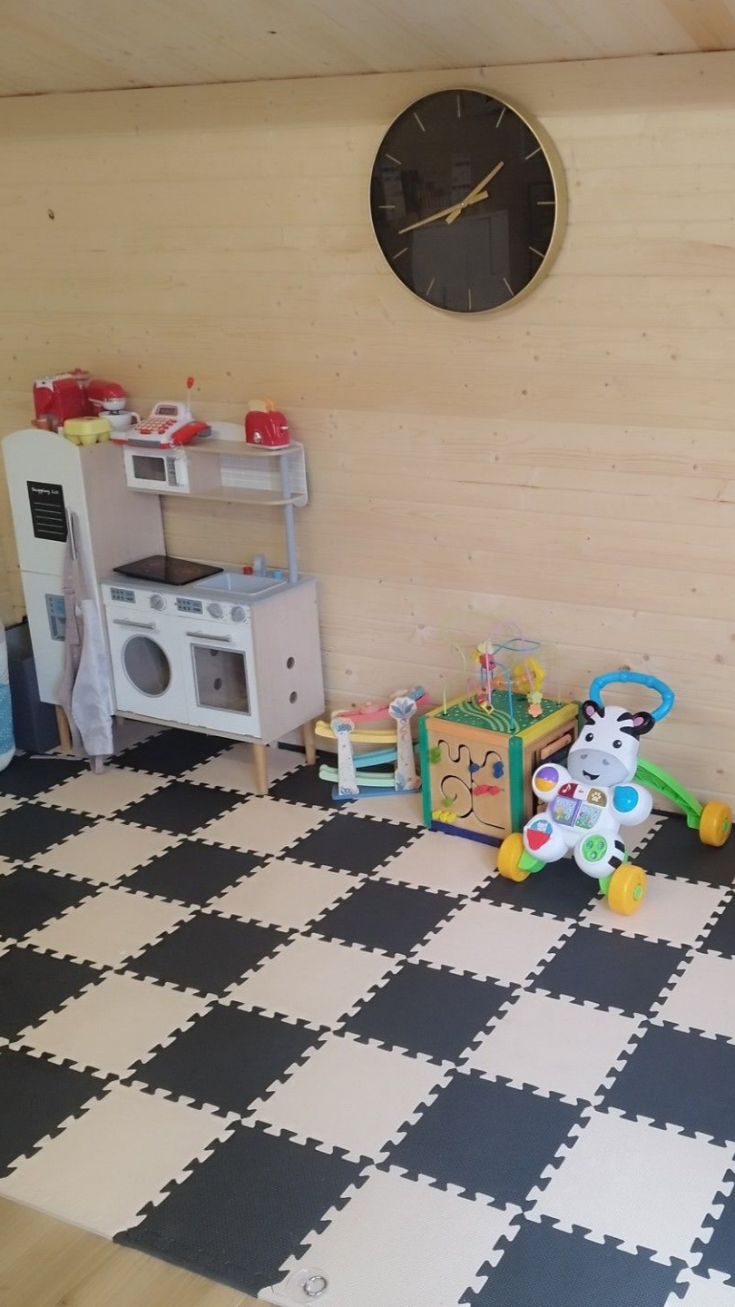 Une aire de jeux pour enfants avec une cuisine jouet, divers jouets, une horloge noire et dorée et un sol à carreaux noir et blanc.