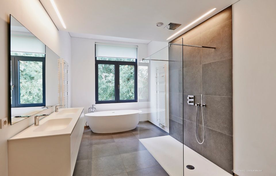 Modernes Badezimmer mit weißem Waschtisch und Badewanne, grau gefliester Dusche und großem Fenster mit Bäumen.