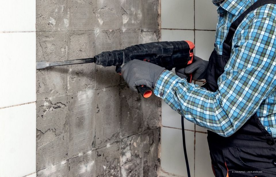 Eine Person in einem blau karierten Hemd benutzt einen Presslufthammer an einer Betonwand.