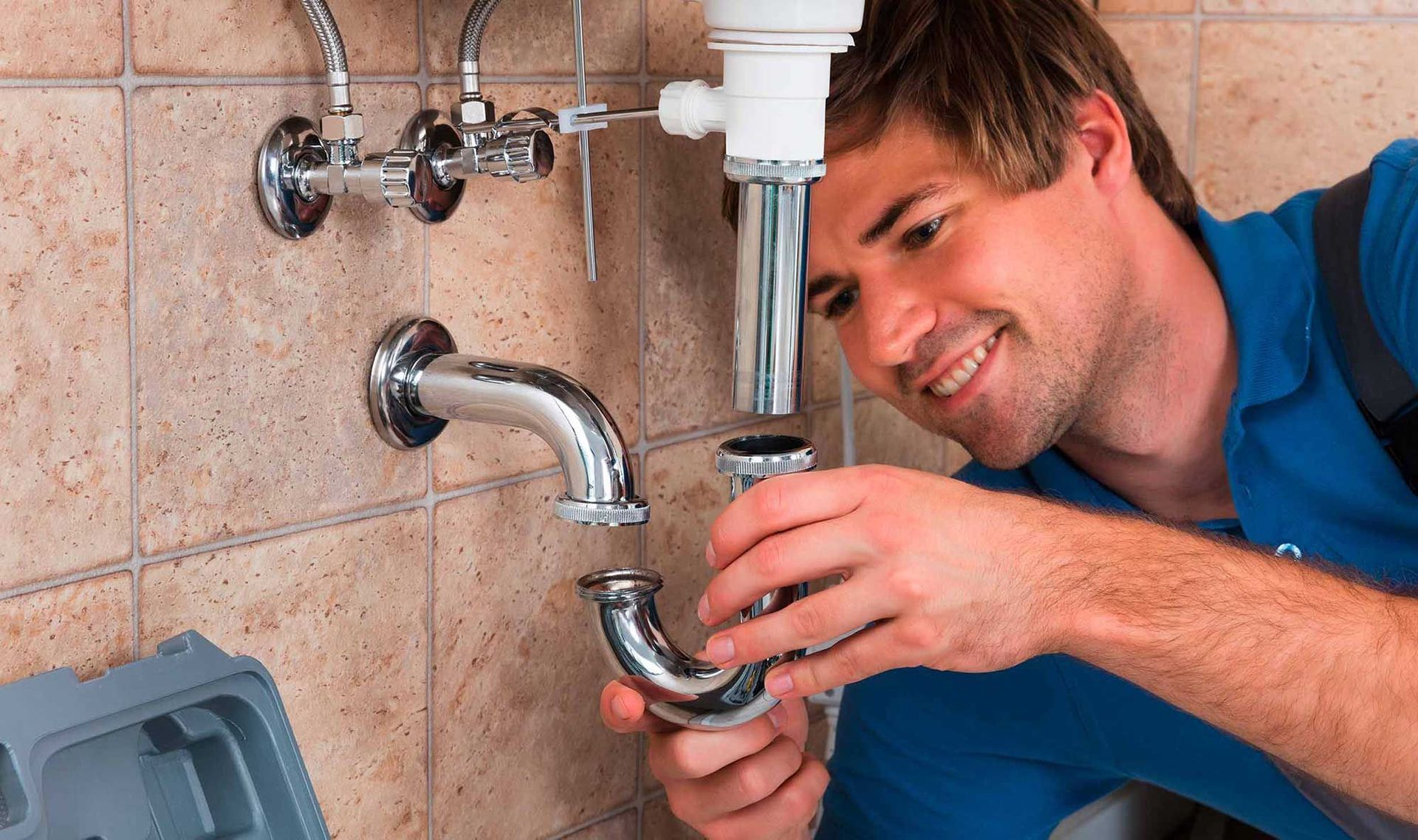 Lächelnder Klempner repariert Rohre unter der Spüle in einem gefliesten Badezimmer.