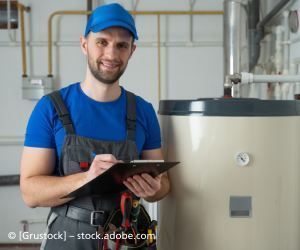 Klempner in blauer Uniform schreibt lächelnd auf ein Klemmbrett neben einem Warmwasserbereiter.
