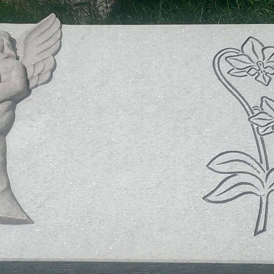 Ein weisser Marmorgrabstein mit einer Engelsdarstellung auf der linken und einem Blumenmotiv auf der rechten Seite.