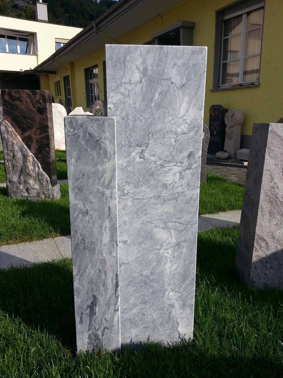 Graue Marmorsäule in einem grasbewachsenen Hof, im Hintergrund weitere Steinbauten und ein gelbes Gebäude.