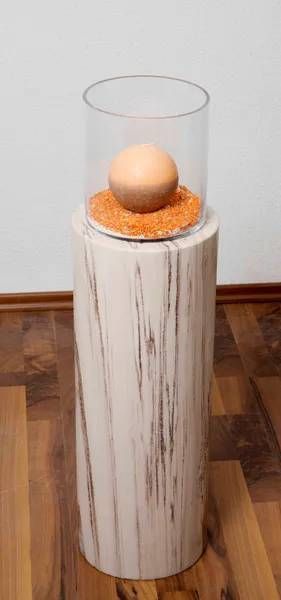 Ein Ei in einem durchsichtigen Zylinder auf einem Bett aus orangefarbenen Körnchen, auf einem strukturierten Holzsockel.