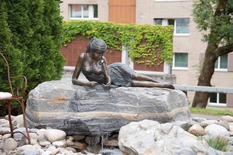 Bronzeskulptur einer Figur, die auf einem grossen Felsen liegt; im Hintergrund sind Grünanlagen und Gebäude zu sehen.