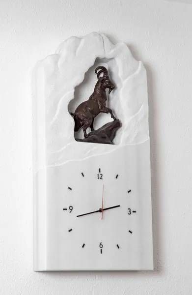Weiße Uhr mit bronzener Bergziegenskulptur.
