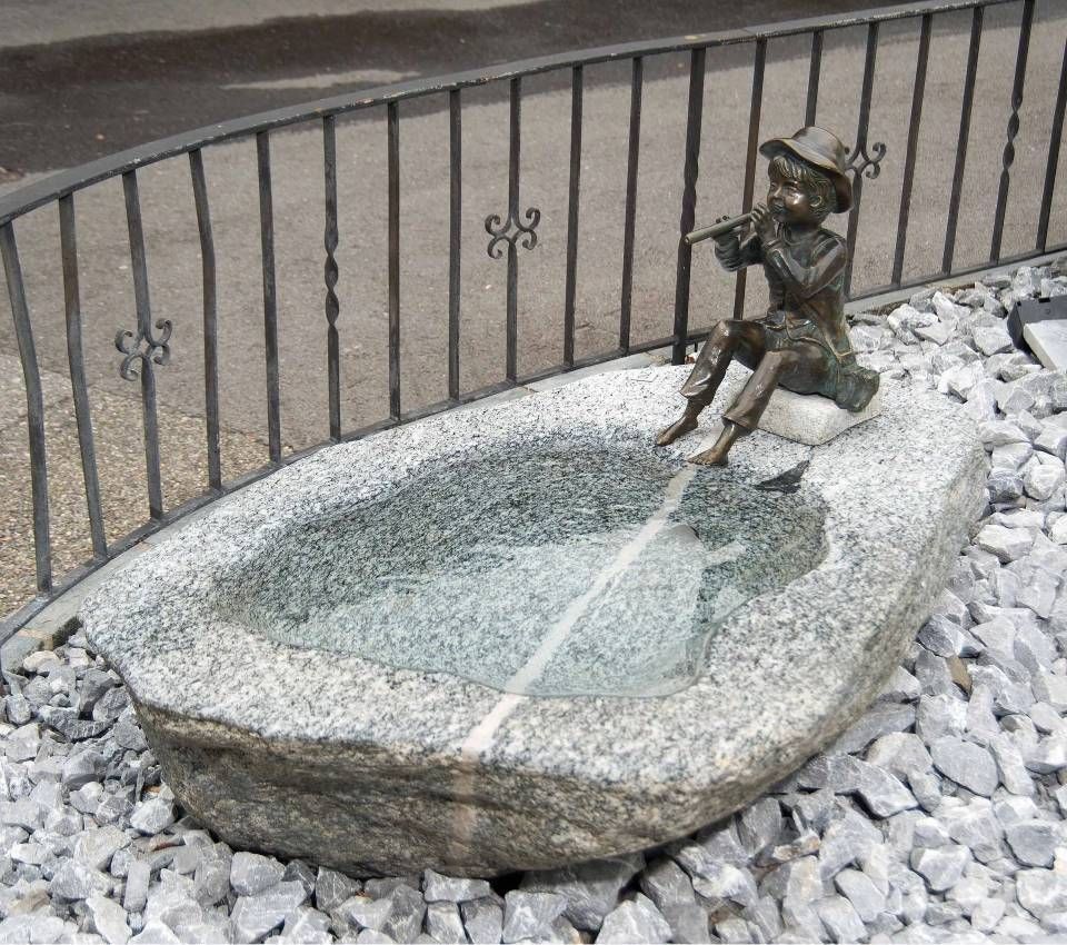 Bronzestatue eines Flöte spielenden Jungen, der auf einem mit Wasser gefüllten Steinbrunnenbecken sitzt und von Kies umgeben ist.