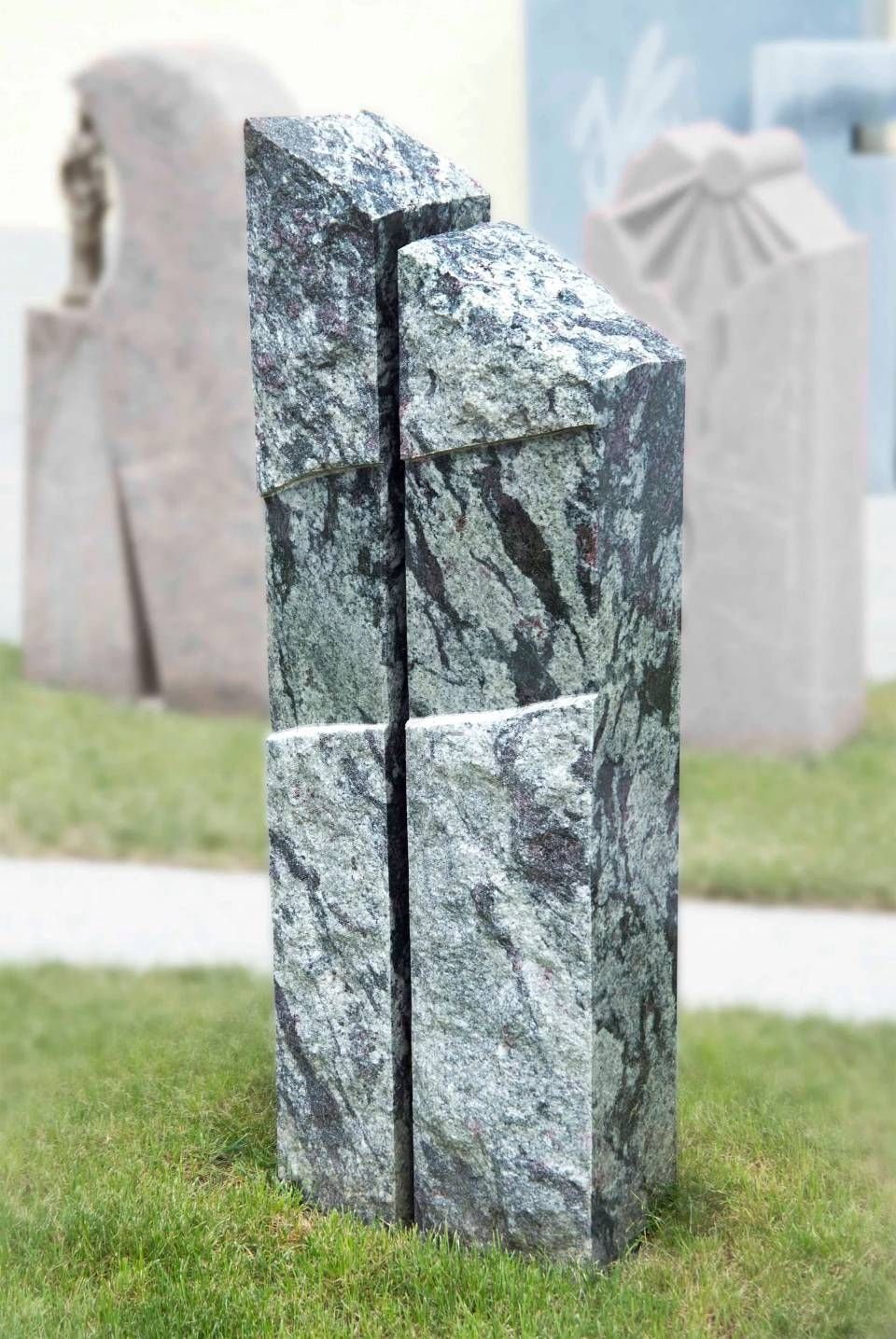 Abstrakte Steinskulptur, möglicherweise ein Denkmal, in einer grasbewachsenen Umgebung. Die Skulptur ist graugrün mit diagonalen Schnittlinien.