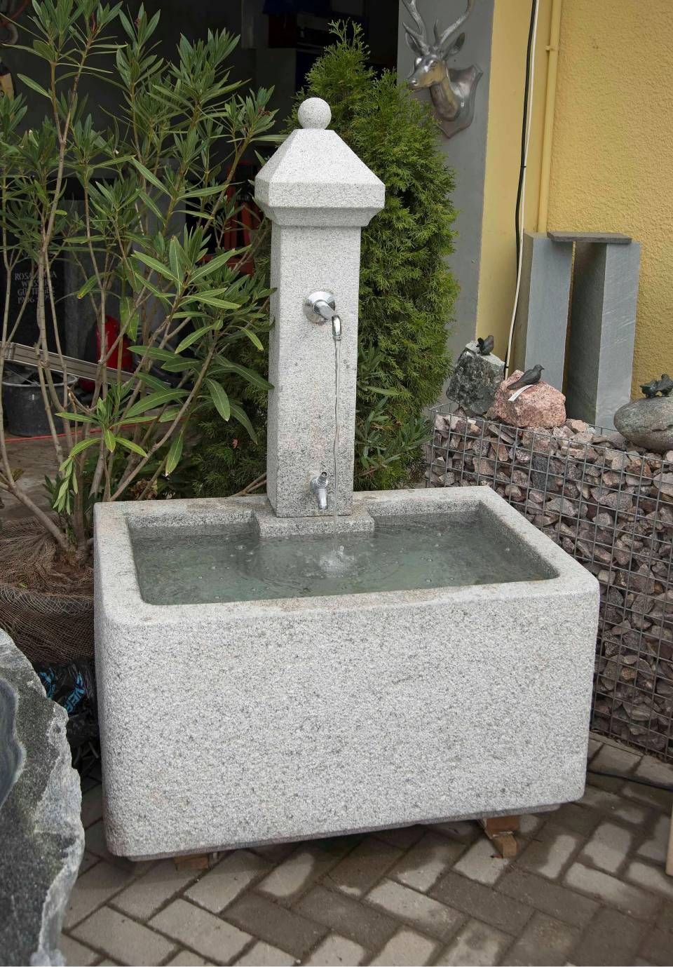 Steinbrunnen mit rechteckigem Becken und hohem, säulenartigem Auslauf. Wasser fliesst aus Hähnen.