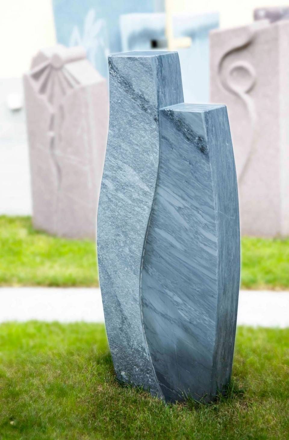Graue Marmorskulptur, zwei miteinander verbundene vertikale Formen, auf grünem Gras; weitere Skulpturen im Hintergrund.