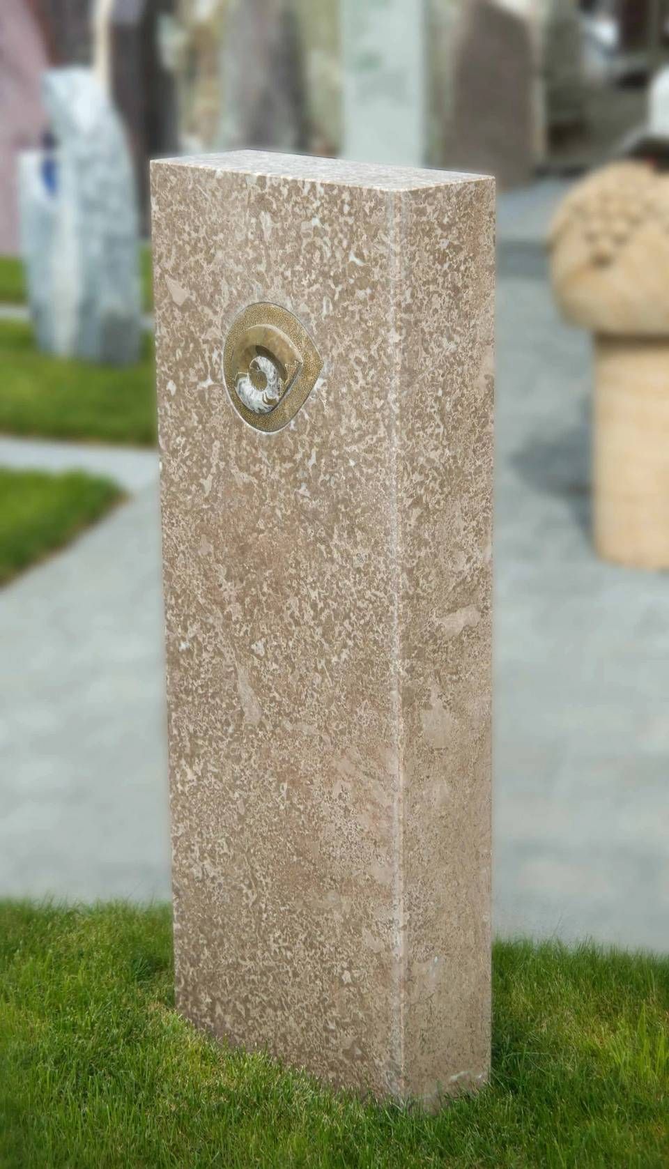 Hohe, rechteckige Steinsäule mit einem Wasserspeier aus Messing in einer grasbewachsenen Aussenanlage.