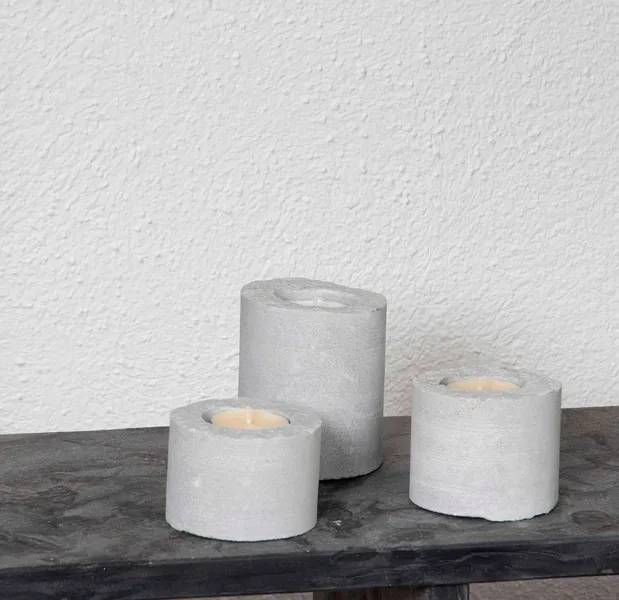 Drei graue Kerzenhalter aus Beton auf dunklem Untergrund, darin brennende Kerzen, vor einer weissen Wand.