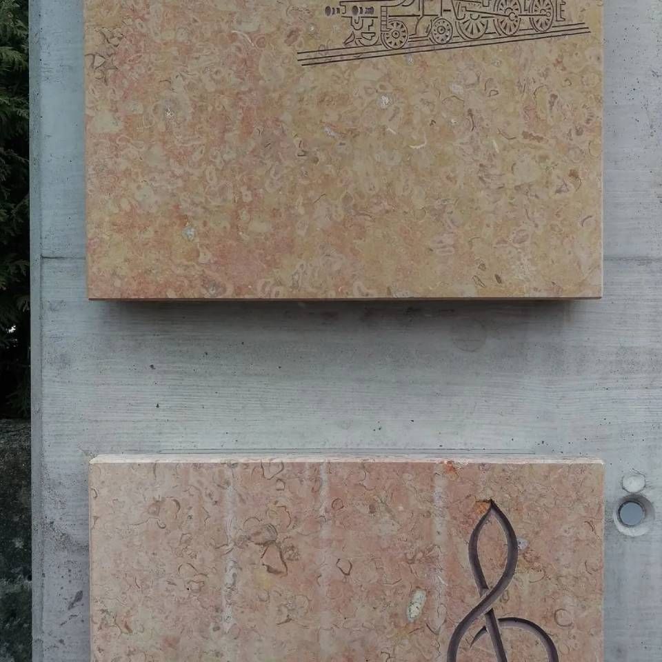 Zwei rechteckige Steintafeln, eine mit einem Zugmotiv, die andere mit einem Violinschlüssel, sind an einer Betonwand angebracht.