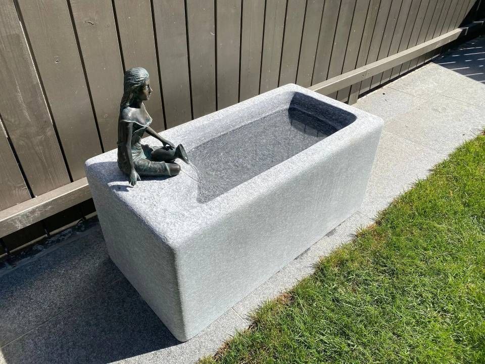 Bronzene Mädchenstatue sitzt am Rand eines rechteckigen grauen Wasserbeckens; im Hintergrund grünes Gras und ein Holzzaun.