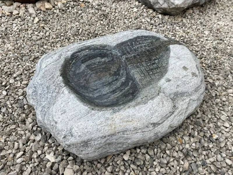 Graues und dunkelgraues Gestein mit kreisförmiger Vertiefung auf einem Kiesbett.