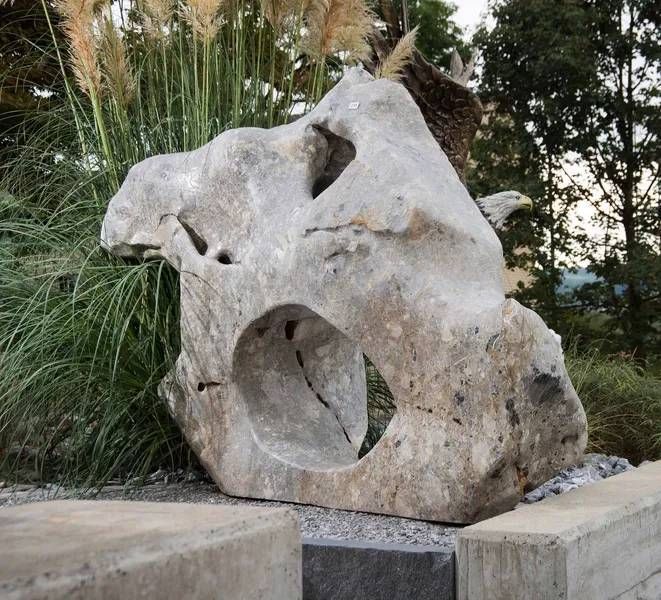Grosse, verwitterte, helle Steinskulptur mit Löchern, im Freien. Hohes Gras und Bäume im Hintergrund.