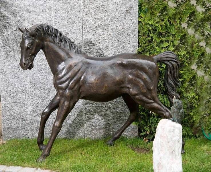 Bronzene Pferdestatue in einer Rasenfläche, vor einer Steinmauer und einer grünen Hecke.