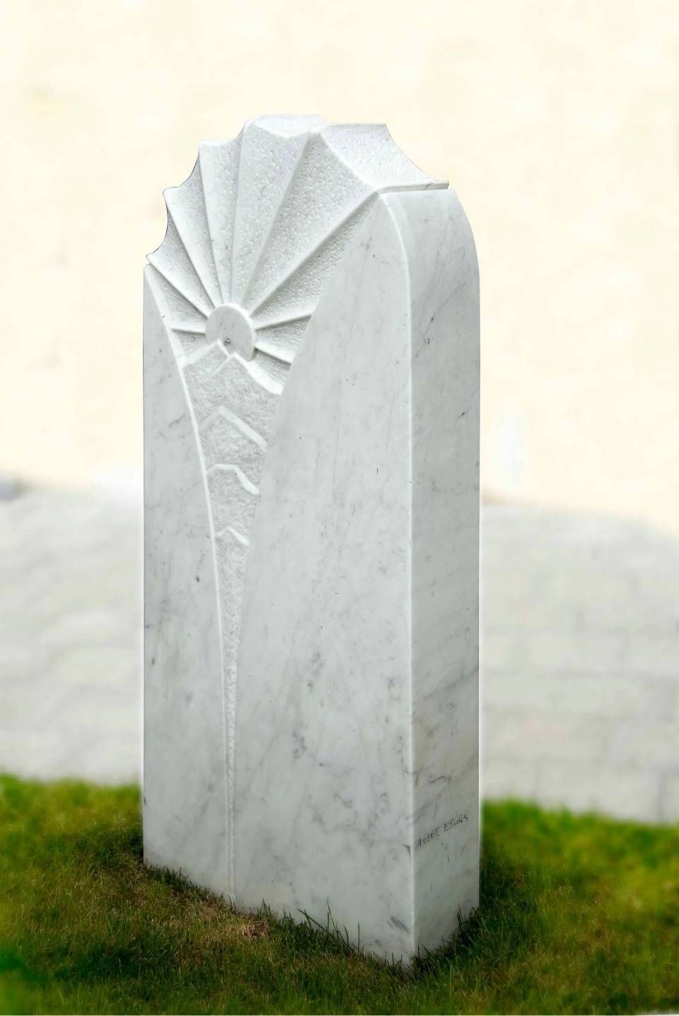 Weisser Marmorgrabstein mit sonnenförmigem Motiv auf grünem Gras.