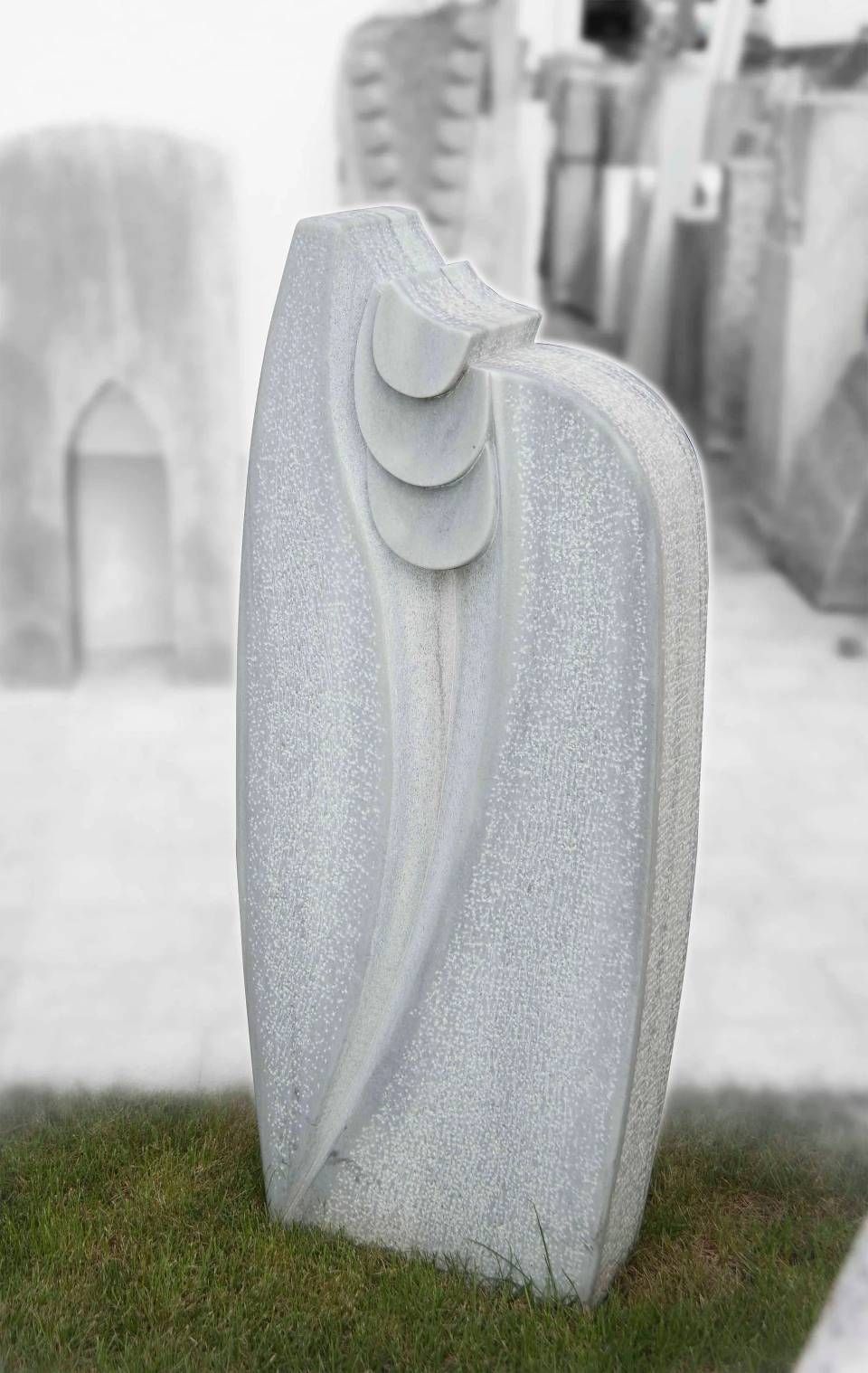 Abstrakte Steinskulptur, hellgrau, mit geschwungenen und geschichteten Details, auf Gras platziert.