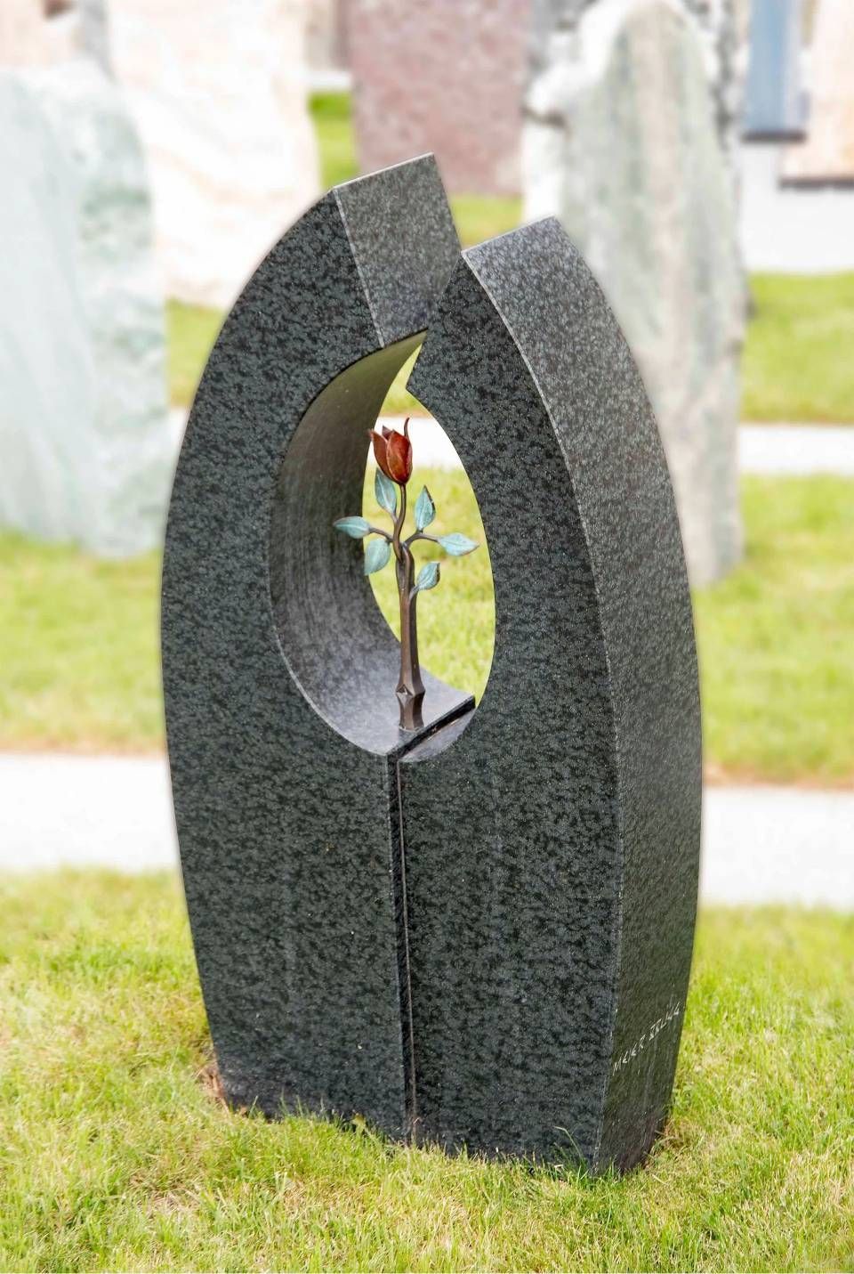 Schwarzes Granitdenkmal mit ovaler Öffnung, darin eine Bronzerose, auf grünem Gras.