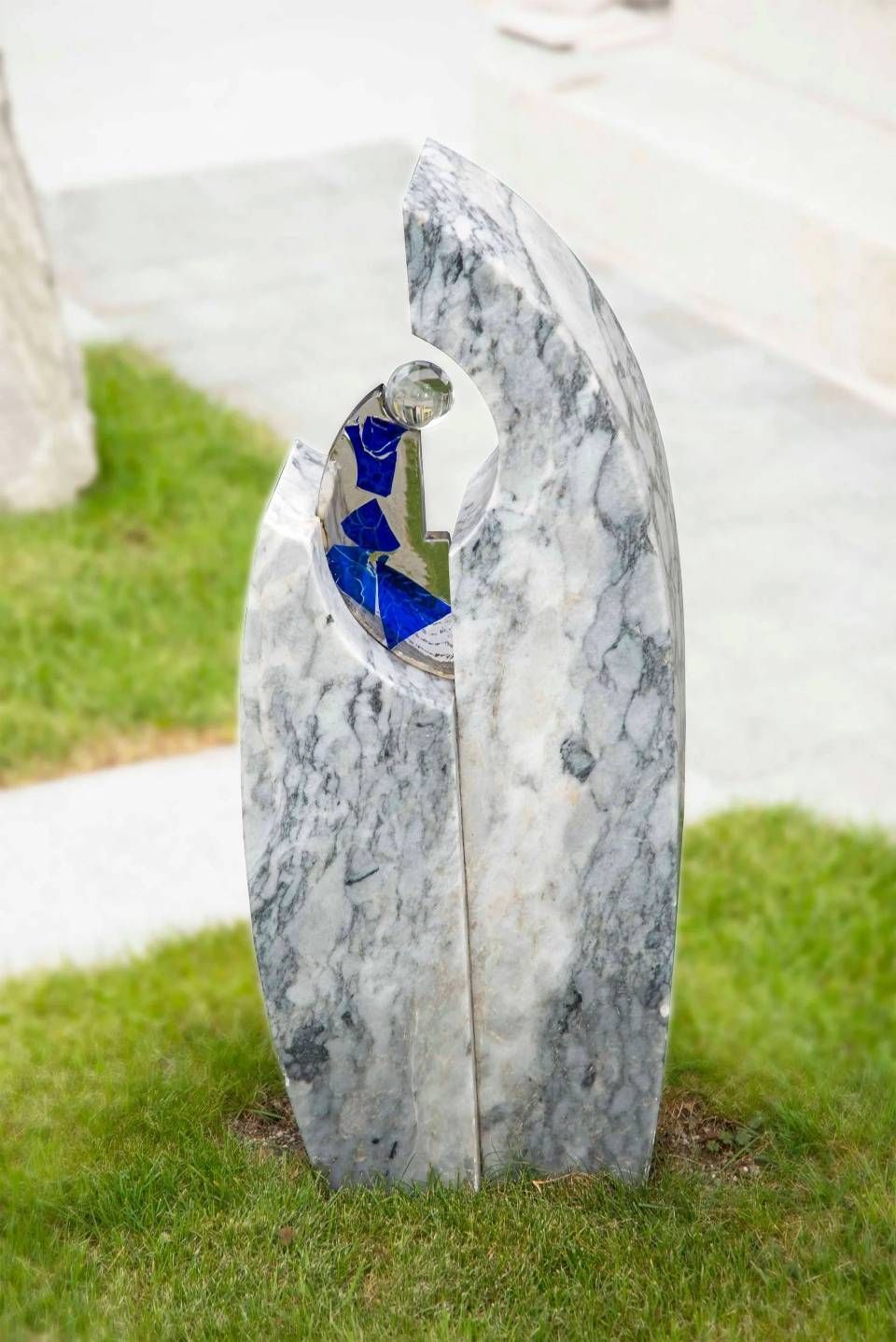 Marmorskulptur einer Mutter mit Kind. Das Kind befindet sich in einer mit blauem Glas gefüllten Vertiefung. Die Skulptur steht auf einer Rasenfläche.