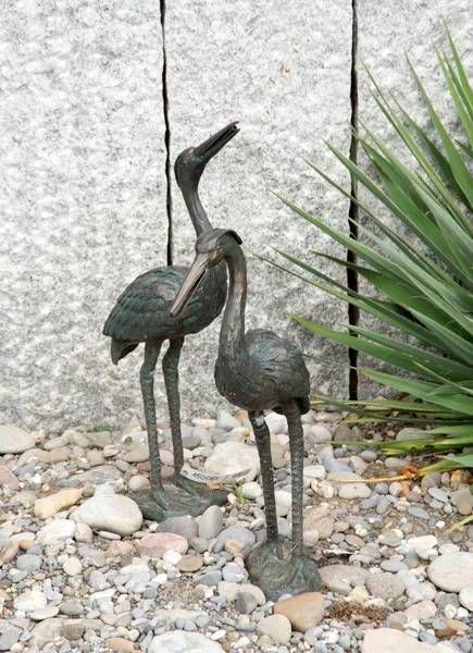 Zwei bronzene Vogelstatuen in einer Gartenanlage, eine blickt nach oben, die andere nach unten, in der Nähe von Felsen und Grünpflanzen.