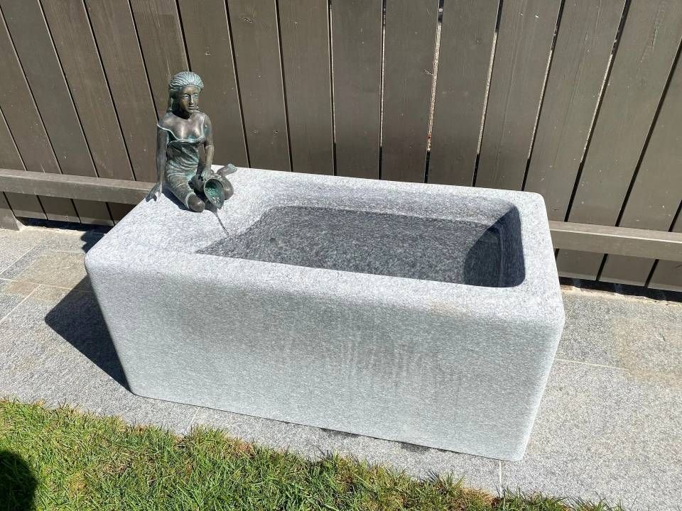 Grauer, rechteckiger Steinbrunnen mit einer kleinen Statue einer sitzenden Frau am Rand.