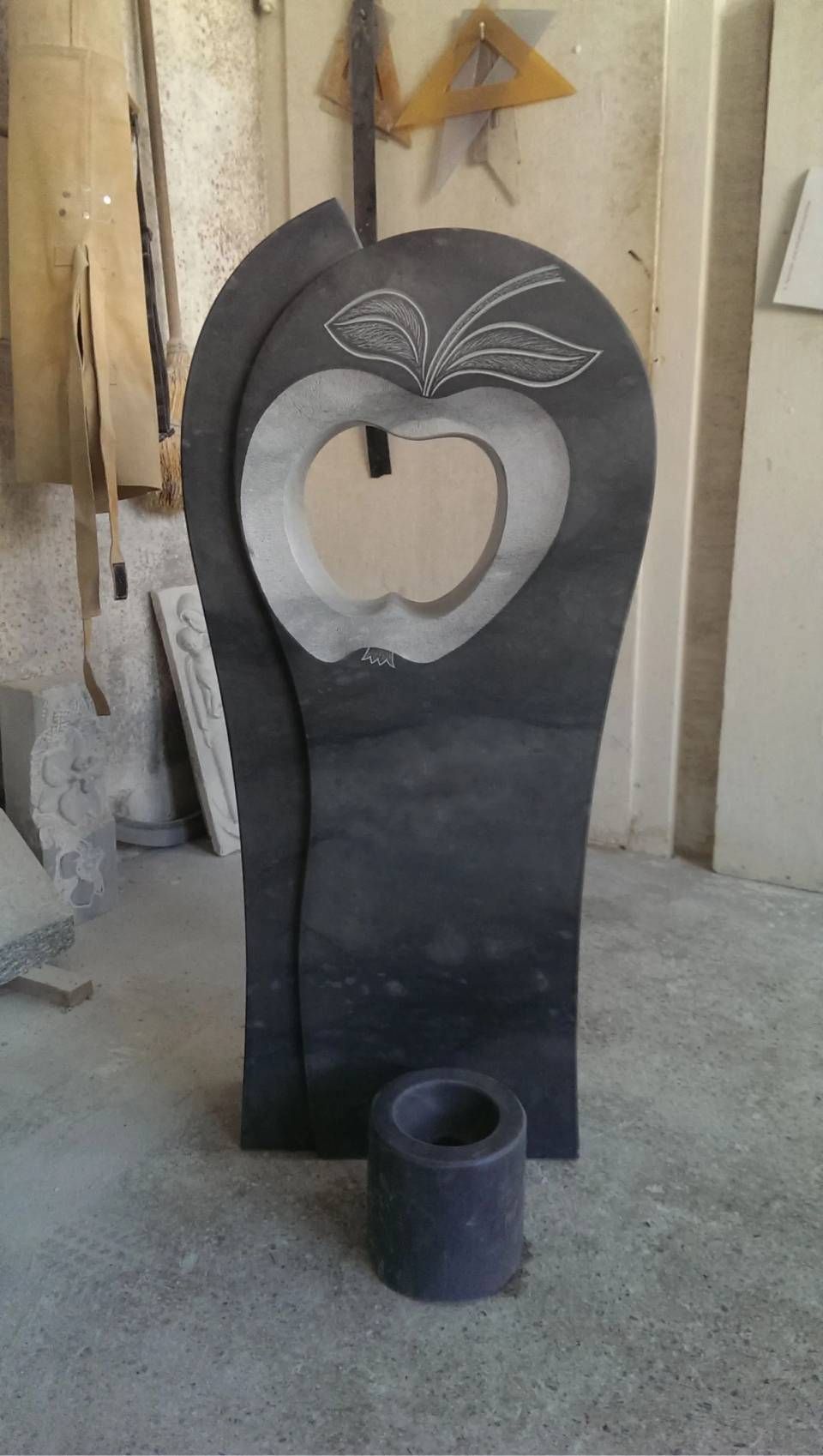 Schwarzer, apfelförmiger Grabstein aus Granit mit ausgeschnittenen und gravierten Details sowie eine kleine dunkle Vase.