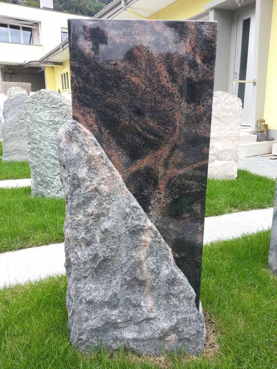 Granitgrabstein mit grob behauenem grauen Stein, abgewinkelt auf schwarzem und braunem poliertem Stein, auf Gras.