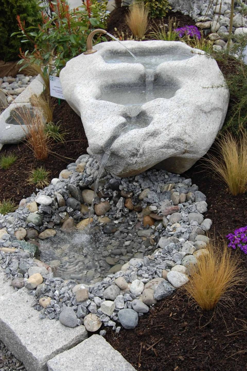 Ein Felsenbrunnen, bei dem das Wasser in ein Steinbett fliesst, das von dunklem Mulch und Pflanzen umgeben ist.