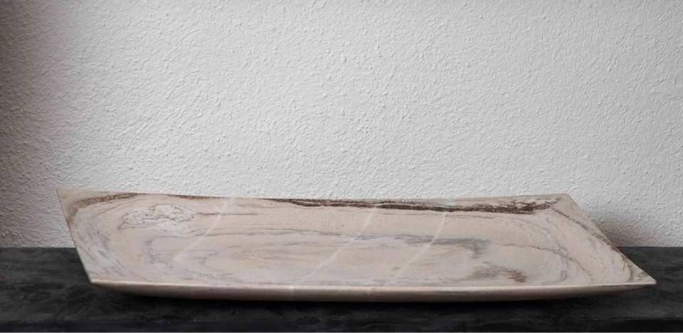 Eine rechteckige Holzschale steht auf einer dunklen Fläche vor einer weissen, strukturierten Wand.