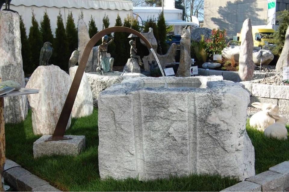 Steinbrunnen, aus dem das Wasser durch einen Metallbogen in ein grob behauenes Becken fliesst; Skulpturen und andere Steinelemente in einer Gartenanlage.