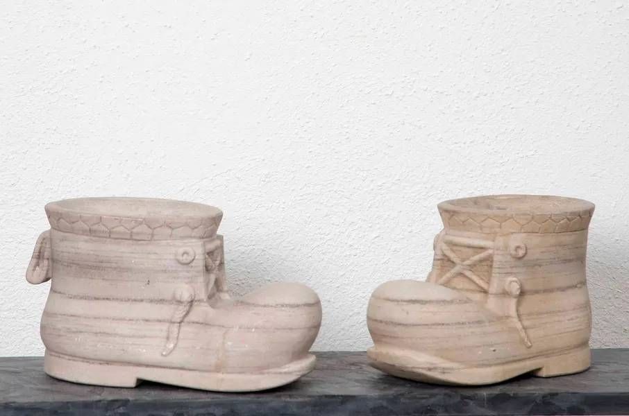Zwei beige, stiefelförmige Pflanzgefässe auf dunklem Untergrund vor einer weissen Wand.
