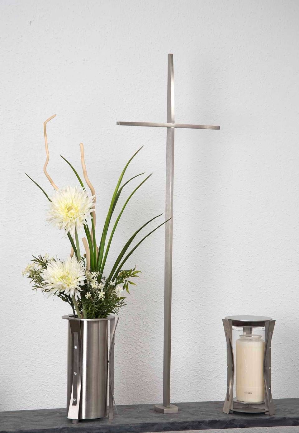 Ein Edelstahlkreuz, eine Vase mit weissen Blumen und ein Kerzenhalter auf einem Regal vor einer weissen Wand.
