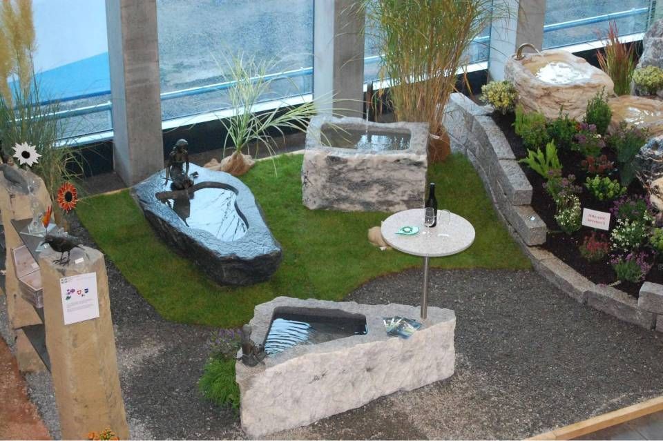Ausstellung von Steinbrunnen und Landschaftsgestaltung in einem Showroom mit Kunstrasen, umgeben von Kies und Grünpflanzen.