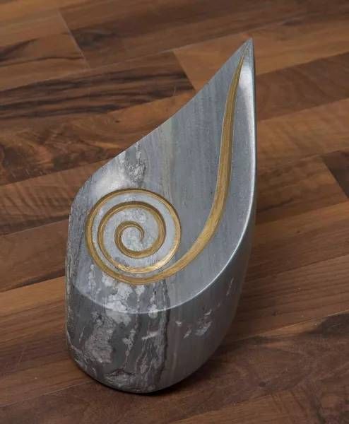 Graue Marmorskulptur mit goldener Spirale und geschwungener Linie auf einem Holzboden.