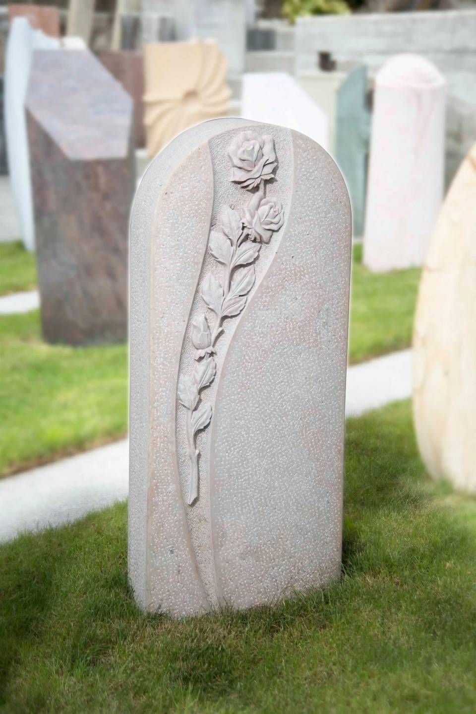 Ein mit Rosen verzierter Grabstein steht auf einer Rasenfläche. Im Hintergrund sind weitere Grabsteine ​​zu sehen.
