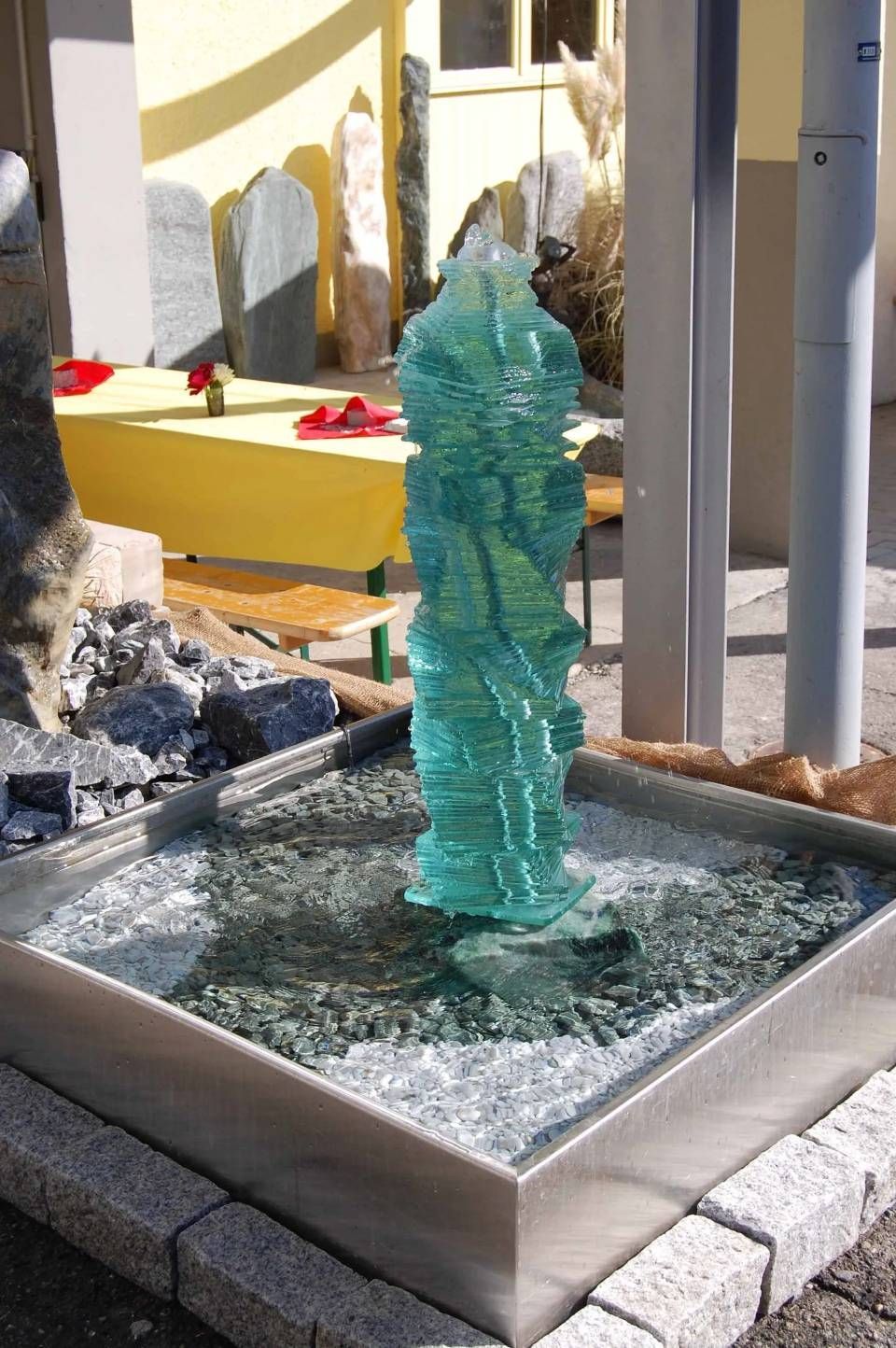 Ein Brunnen aus türkisfarbenem Glas in einem Edelstahlbecken, umgeben von Steinen.
