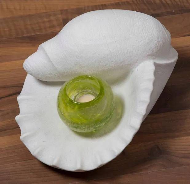 Weisser, muschelförmiger Kerzenhalter mit einem grünen Glasvotiv und einer brennenden Kerze auf einer Holzoberfläche.