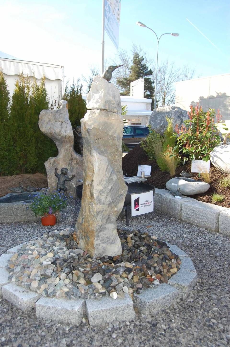 Steinbrunnen mit hohen, strukturierten Felsen in einem kreisförmigen Steinbett, umgeben von Kies.