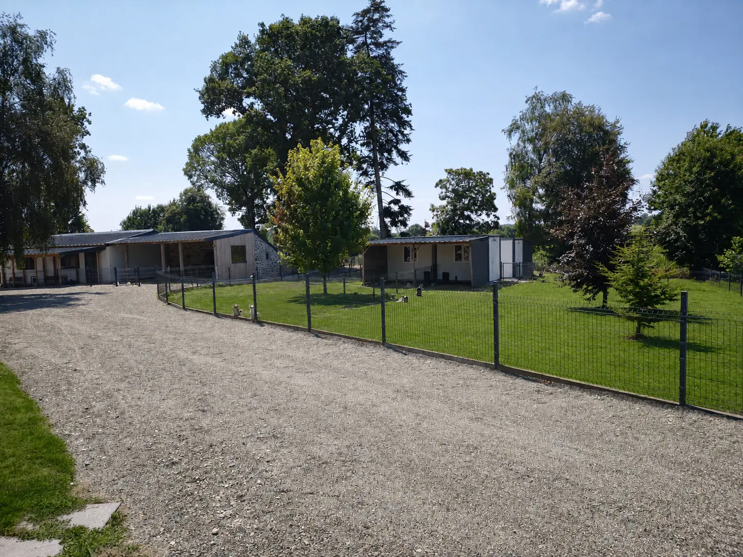 Allée de gravier longeant une pelouse verte clôturée, agrémentée de petits bâtiments.
