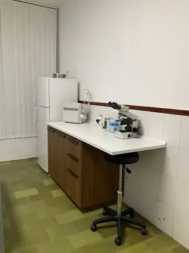 Pequeña zona de cocina con encimera blanca, mininevera, cafetera y taburete junto a una pared alicatada.