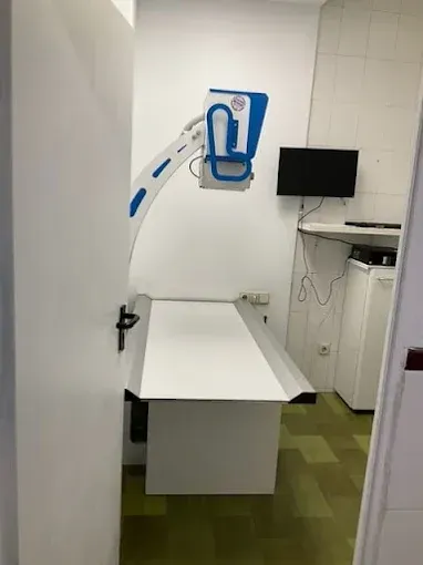 Pequeña sala de exploración médica con una camilla blanca, equipo montado en la pared y un monitor.