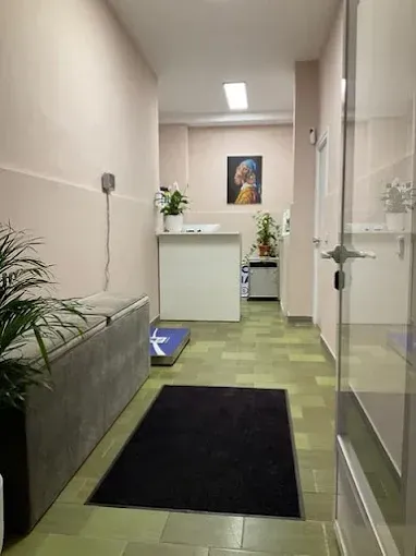 Pasillo estrecho con alfombra negra, banco gris, puerta de cristal y un pequeño escritorio al fondo.