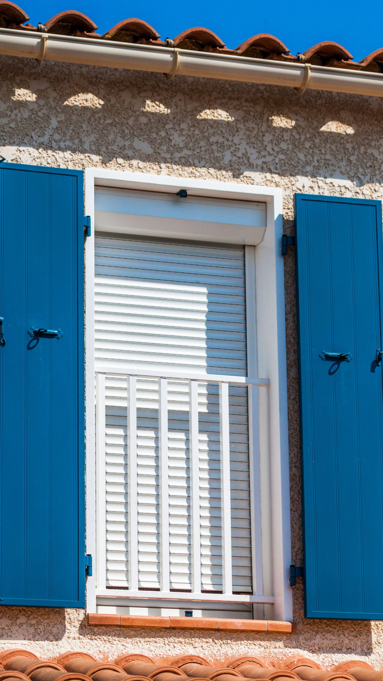 Vue depuis l'extérieur d'un volet roulant en PVC installé sur une fenêtre équipée de volets battants en bois bleu