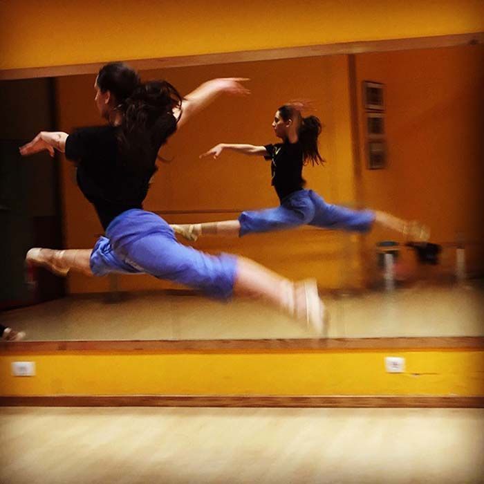 Dos bailarines con pantalones azules saltan por un estudio con espejos y paredes de color naranja cálido.