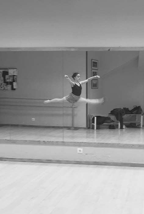 Bailarina de ballet con leotardo negro haciendo un salto con apertura de piernas frente a un espejo en la barra del estudio.