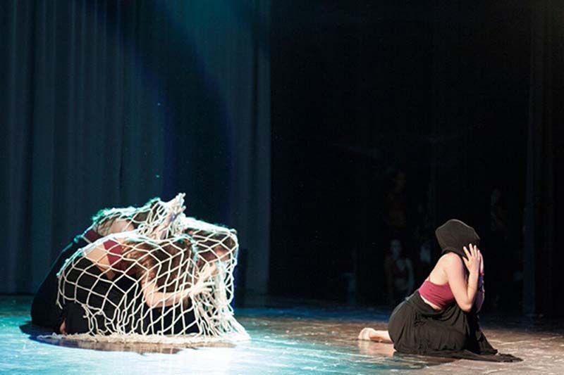 La bailarina se arrodilla en el escenario junto a una red blanca enredada que cubre un objeto bajo, sobre un fondo oscuro.