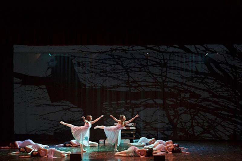 Bailarinas de ballet con tutús blancos posan en el escenario entre varias bailarinas tumbadas en un decorado oscuro y sombrío.