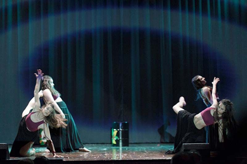 Dos bailarinas realizando posturas simétricas de flexión hacia atrás sobre un escenario iluminado con un telón oscuro de fondo.
